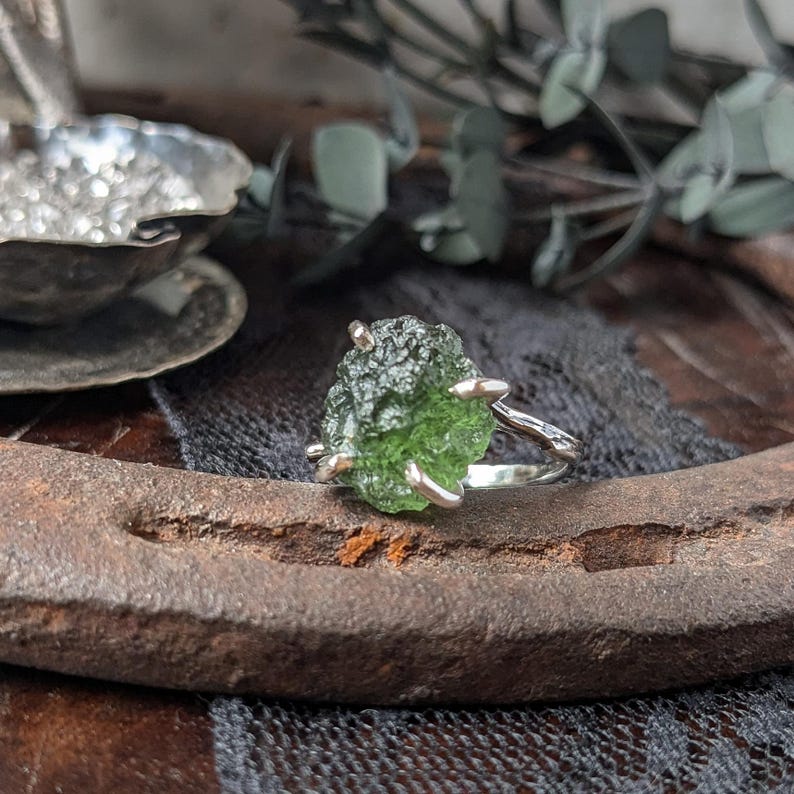 May include: A silver ring with a green, rough, natural gemstone. The ring is set on a rustic, brown surface.