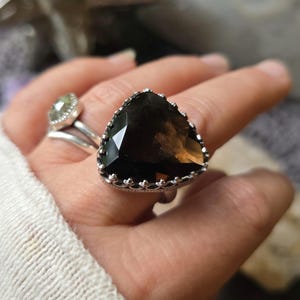 May include: A large, triangular-cut smoky quartz gemstone ring with a silver crown setting. The dark brown stone has multiple facets. Another ring with a smaller, light green stone is also visible.