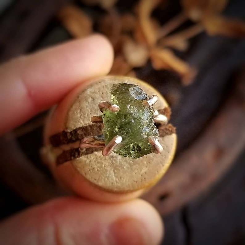 May include: A silver ring with a raw, green peridot gemstone set in a claw setting. The ring is on a wooden display stand.