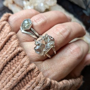 May include: A silver ring with a clear quartz crystal set in a wire wrap design. The ring is worn on a hand with a silver ring with a green gemstone.