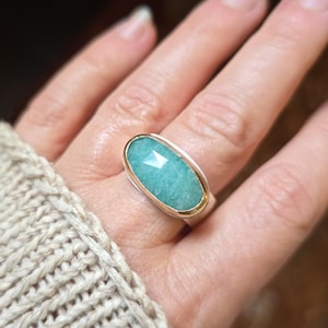 Puede incluir: Un anillo de plata con una gema grande, ovalada y azul claro. La gema está facetada y engastada en un bisel dorado, con una banda de plata. El anillo está en el dedo de una persona, con un suéter beige en el fondo.