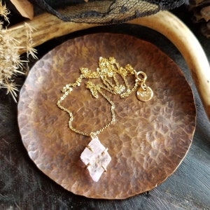 Pu&ograve; includere: Una delicata collana a catena dorata con un ciondolo di cristallo rosa. Il ciondolo &egrave; una forma geometrica sfaccettata con una montatura in oro. La collana &egrave; esposta su un piatto di rame martellato.