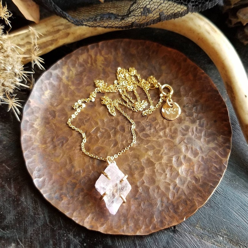 Pu&ograve; includere: Una delicata collana a catena dorata con un ciondolo di cristallo rosa. Il ciondolo &egrave; una forma geometrica sfaccettata con una montatura in oro. La collana &egrave; esposta su un piatto di rame martellato.