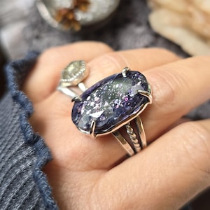Puede incluir: Un anillo de plata con una gran piedra ovalada de color morado intenso con inclusiones brillantes plateadas y rosas. La piedra está sujeta por cuatro garras en una banda que incluye un detalle de metal trenzado. Se ve un segundo anillo de plata detrás.