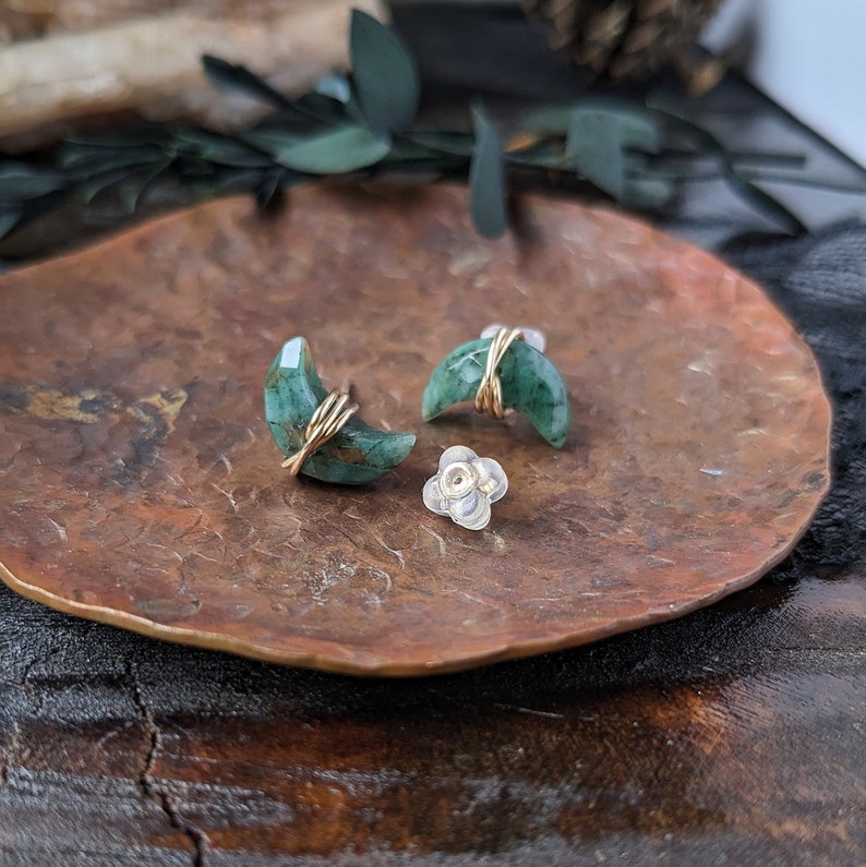 May include: A pair of crescent moon shaped earrings made with green gemstone and gold wire. The earrings are on a copper-colored plate.
