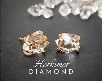 May include: A pair of gold earrings featuring raw Herkimer diamond crystals. The diamonds are clear and have a natural, rough texture. The earrings are a simple and elegant design.