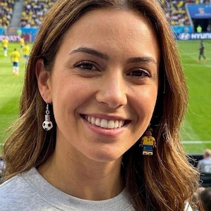 May include: A woman wearing soccer-themed earrings. One earring is a soccer ball, the other is a Lego figure wearing a yellow and blue soccer uniform. The background shows a soccer field with players in yellow uniforms.