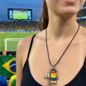 May include: A black cord necklace with a pendant featuring a soccer player figurine and a soccer ball charm. The player wears a yellow and blue uniform. The background shows a soccer stadium with a large screen.