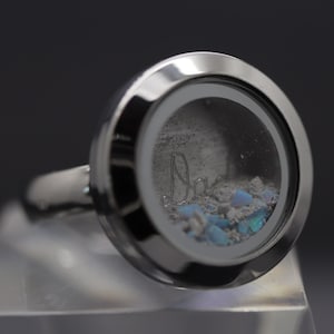 Può includere: Un anello d'argento con un ciondolo di vetro trasparente. Il ciondolo contiene glitter blu e bianco e la parola "Papà" è incisa all'interno.