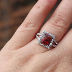 May include: A silver ring with a square-shaped red gemstone surrounded by smaller clear gemstones.
