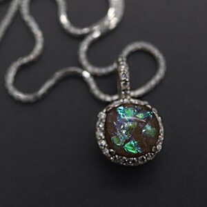 May include: A silver chain necklace with a round pendant featuring a dark brown stone with green and blue iridescent glitter accents. The pendant is surrounded by a silver bezel with small clear stones.