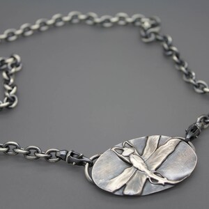 May include: A silver chain necklace with a silver oval pendant featuring a dragonfly design.