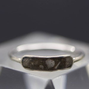 May include: A silver ring with a rectangular, dark gray stone inset. The stone has a speckled appearance with lighter gray and white inclusions. The ring is displayed on a clear, square block against a dark background.