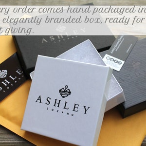 May include: Three gift boxes with the logo "Ashley Lozano" printed on them. The boxes are white, black, and gray. The boxes are on a brown wooden surface.
