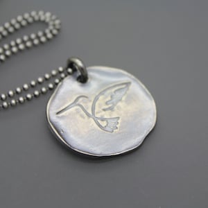 Op de afbeelding: Een zilveren ketting met een ronde hanger met een gegraveerd kolibrie ontwerp.
