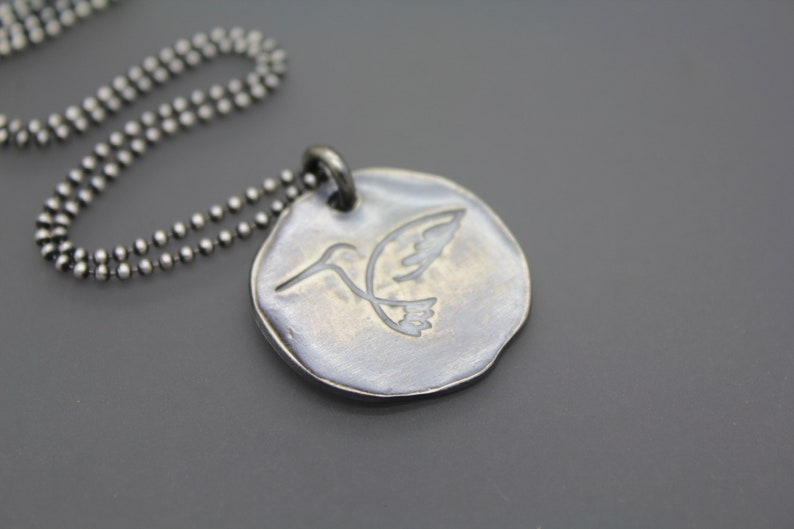 Op de afbeelding: Een zilveren ketting met een ronde hanger met een gegraveerd kolibrie ontwerp.