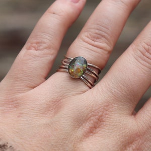 Op de afbeelding: Een zilveren ring met een veelkleurige ovale steen in het midden. De ring heeft drie banden die om de vinger wikkelen.