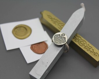 Può includere: Un anello in argento con una faccia circolare che presenta un design inciso di montagna e sole, appoggiato su un bastoncino di cera argentato. Due sigilli in cera, uno dorato e uno color rame, sono posti su quadrati di carta bianca vicino a due bastoncini di cera dorati.