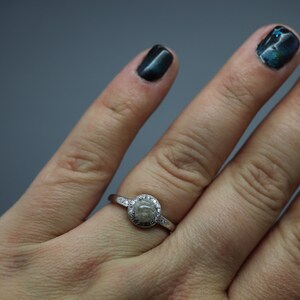 May include: A silver ring featuring a round, grey stone encircled by smaller clear stones. The ring is worn on a person's finger.