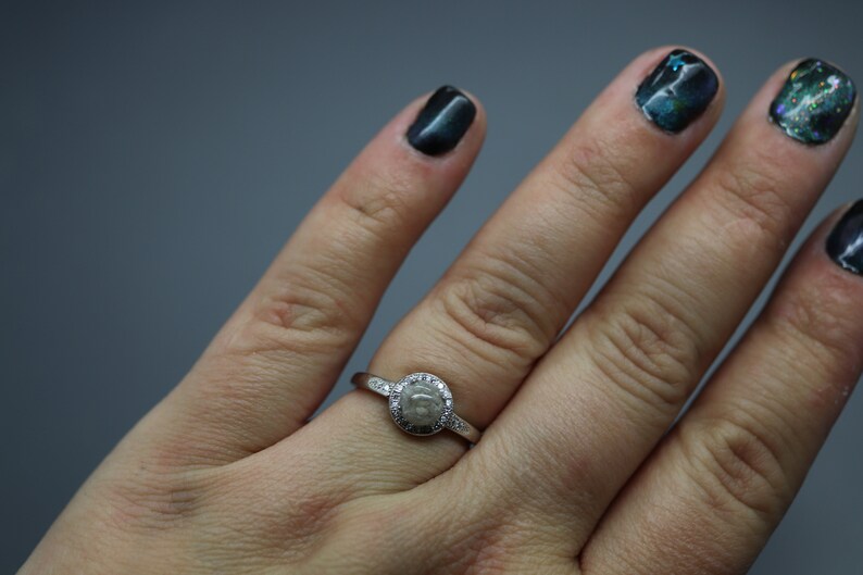May include: A silver ring featuring a round, grey stone encircled by smaller clear stones. The ring is worn on a person's finger.