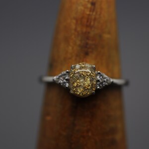 May include: A silver ring with a clear oval-shaped stone filled with gold flakes. The stone is set in a silver band with two smaller clear stones on either side.
