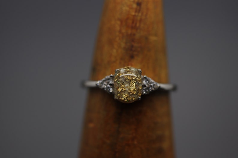 May include: A silver ring with a clear oval-shaped stone filled with gold flakes. The stone is set in a silver band with two smaller clear stones on either side.