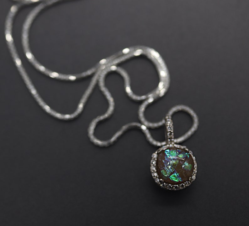 May include: A silver chain necklace with a round pendant featuring a dark brown stone with green and blue iridescent glitter accents. The pendant is surrounded by a silver bezel with small clear stones.
