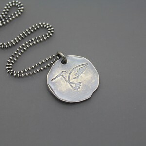 Op de afbeelding: Een zilveren ketting met een ronde hanger met een gegraveerd kolibrie-ontwerp.
