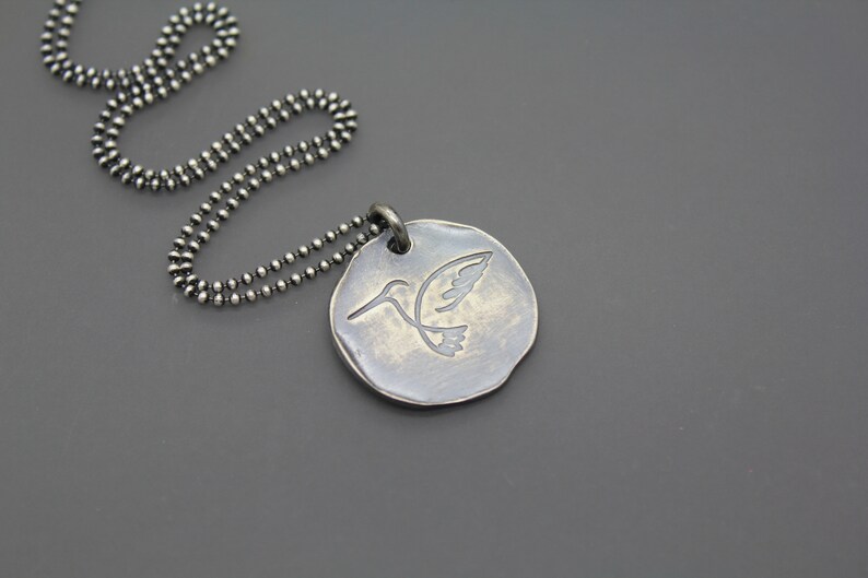 Op de afbeelding: Een zilveren ketting met een ronde hanger met een gegraveerd kolibrie-ontwerp.