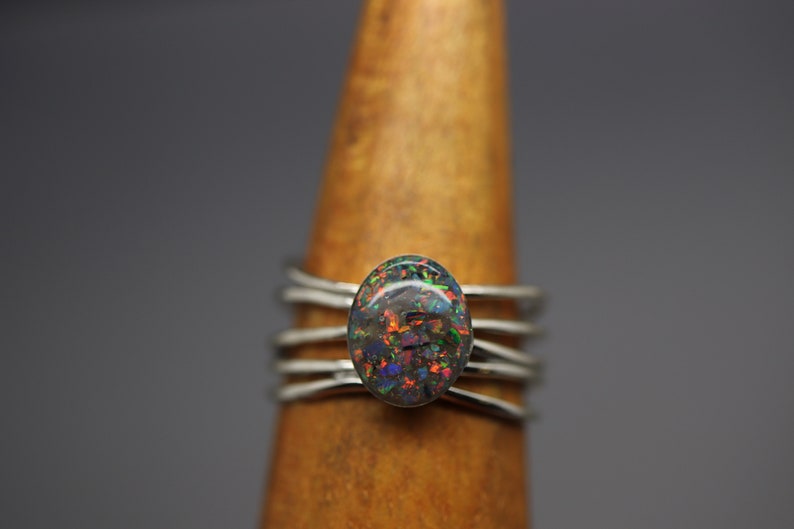 Op de afbeelding: Een zilveren ring met een veelkleurige opaal edelsteen in het midden. De ring heeft een eenvoudig, modern ontwerp met meerdere banden.