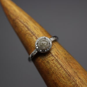 May include: A silver ring featuring a round, grey stone set within a halo of small diamonds.