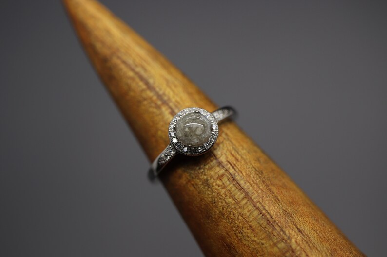 May include: A silver ring featuring a round, grey stone set within a halo of small diamonds.