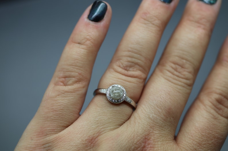 May include: A silver ring featuring a round, clear stone set within a halo of smaller stones. The ring is worn on a finger.