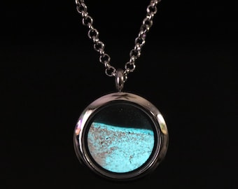 Peut inclure: Un pendentif médaillon circulaire en argent suspendu à une chaîne en argent assortie. Le médaillon présente une face en verre transparent révélant une surface intérieure texturée bleu clair et sable rappelant un rivage.