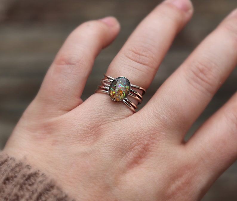Op de afbeelding: Een zilveren ring met een veelkleurige, ovale steen in het midden. De ring heeft drie banden die om de vinger wikkelen.
