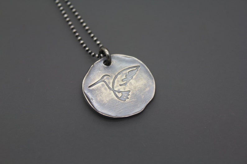 Op de afbeelding: Een zilveren ketting met een ronde hanger met een kolibrie-ontwerp.