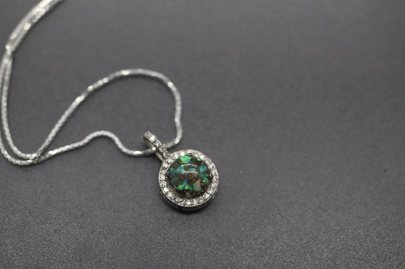 May include: A silver chain necklace with a round pendant featuring a green and black opal stone surrounded by a halo of clear cubic zirconia stones.