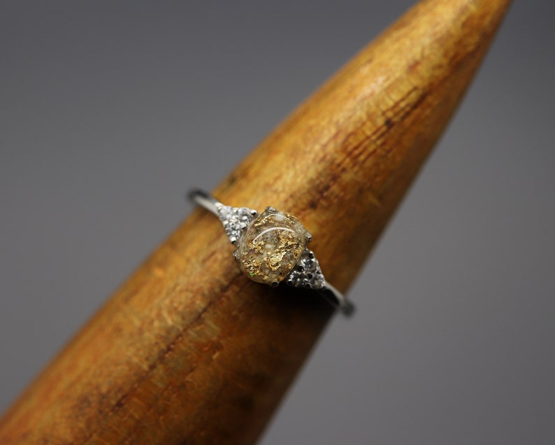 May include: A silver ring with a clear stone set in the center. The stone is filled with small pieces of gold leaf. The ring is set on a wooden display stand.