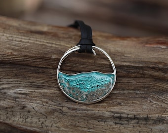 Può includere: Un ciondolo circolare in argento con un design a onda turchese testurizzato sopra uno strato di materiale granulare simile alla sabbia. Il ciondolo è sospeso a un cordino nero, creando un contrasto tra la cornice metallica e gli elementi.