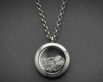 Peut inclure: Un pendentif médaillon circulaire de couleur argentée suspendu à une chaîne maille forçat assortie. Le médaillon présente une face en verre transparent révélant une collection de petits fragments texturés multicolores et des pierres claires.