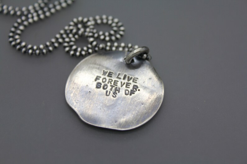 Op de afbeelding: Zilveren ketting met een ronde hanger met de tekst "We leven voor altijd. Beide van ons."