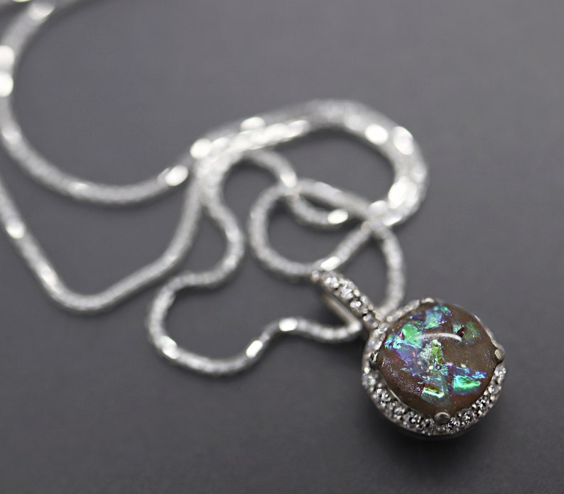 K&ouml;nnte beinhalten: Eine silberne Kettenkette mit einem Anh&auml;nger mit einem quadratischen, regenbogenfarbenen Opal-Stein, der in einer silbernen Fassung mit einer Reihe kleiner, klarer Steine um den Rand herum gefasst ist.
