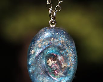 May include: An oval-shaped pendant on a silver-toned chain. The pendant features a swirling, iridescent blue and purple resin surface embedded with fine glitter and small, light-colored granular particles throughout the center.