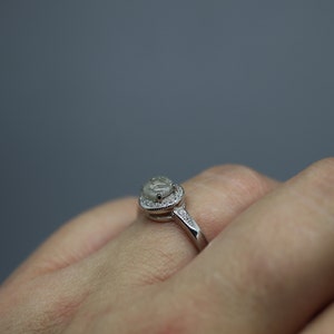 May include: A silver ring featuring a large, clear, oval-shaped gemstone encircled by a halo of smaller gemstones.