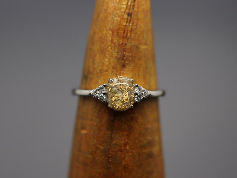 May include: A silver ring with a clear oval-shaped stone filled with gold flakes. The stone is surrounded by two smaller clear stones.