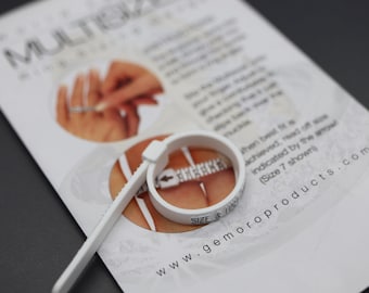 May include: A white plastic ring sizer tool with a buckle and a printed scale, resting on an instructional card labeled MULTISIZER and gemoroproducts.com. The tool features a loop with a black arrow indicator and the text SIZE & HALF SIZES.
