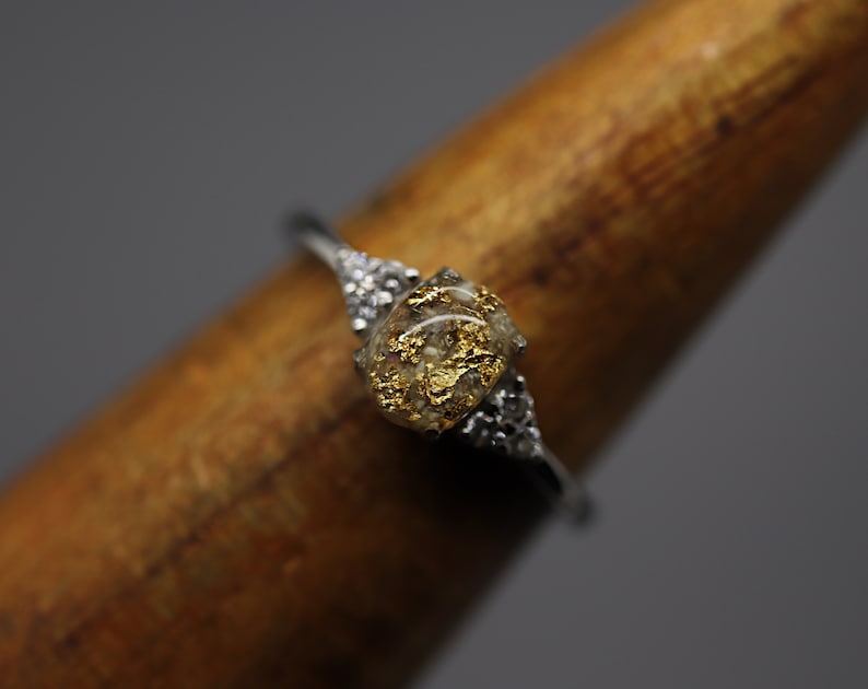 May include: A silver ring with a square-shaped clear stone filled with gold flakes. The stone is set in a silver band with small silver accents.