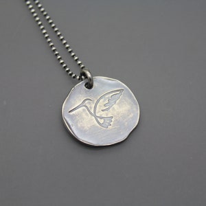 Op de afbeelding: Een zilveren ketting met een ronde hanger met een kolibrie-ontwerp. De hanger heeft een getextureerd oppervlak en een zilveren ketting.