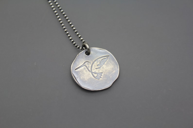 Op de afbeelding: Een zilveren ketting met een ronde hanger met een kolibrie-ontwerp. De hanger heeft een getextureerd oppervlak en een zilveren ketting.