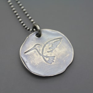 Op de afbeelding: Een zilveren ketting met een ronde hanger met een kolibrie-ontwerp. De hanger heeft een getextureerde, verweerde afwerking.